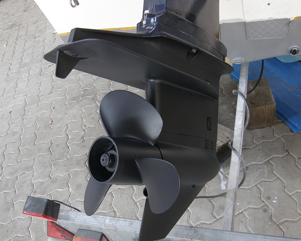 Black three-blade propeller and lower gearcase of an outboard motor beside a boat trailer.