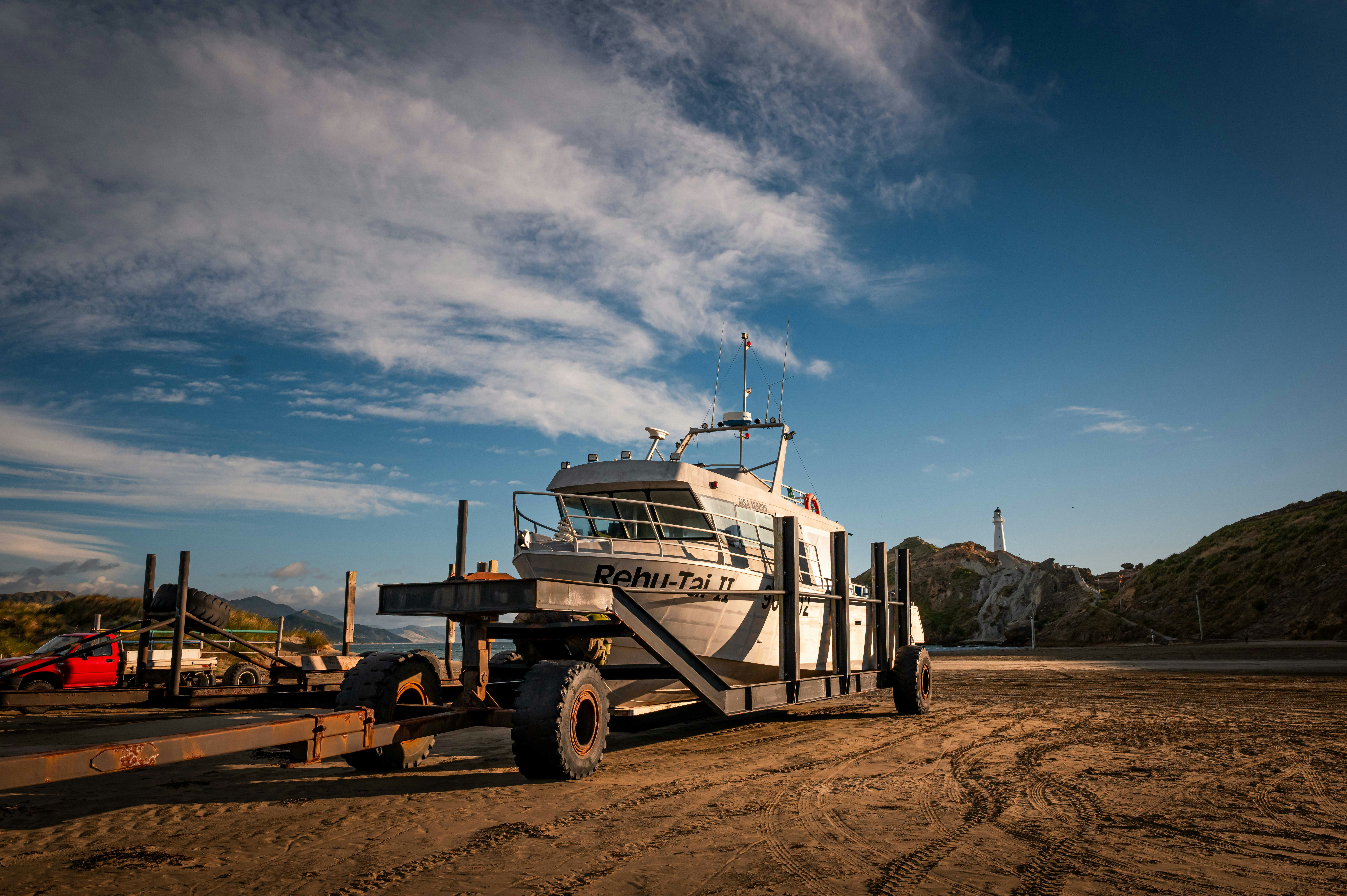 A white cabin fishing boat rests ashore on a wheeled trailer beneath a cloudy blue sky.